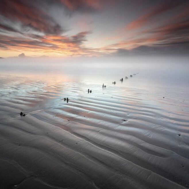 rippled tidal sand at dusk, a line of worn posts receding into sea mist.